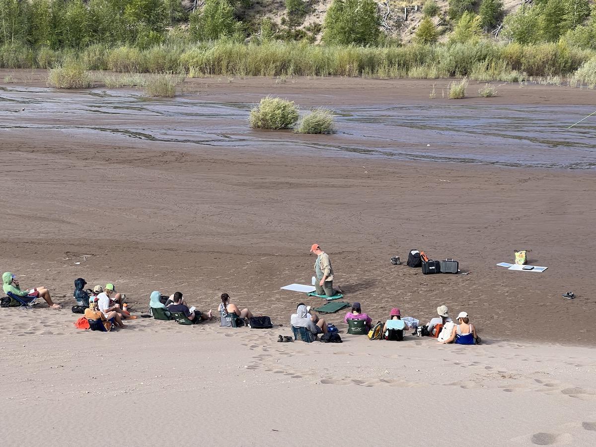 Class of students sitting on sand with a stream behind them studying a riparian zone